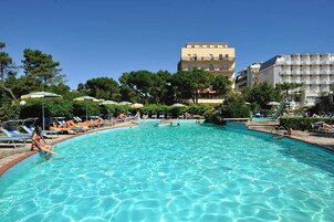 Una piscina con lettini e ombrelloni, circondata da alberi ed edifici.