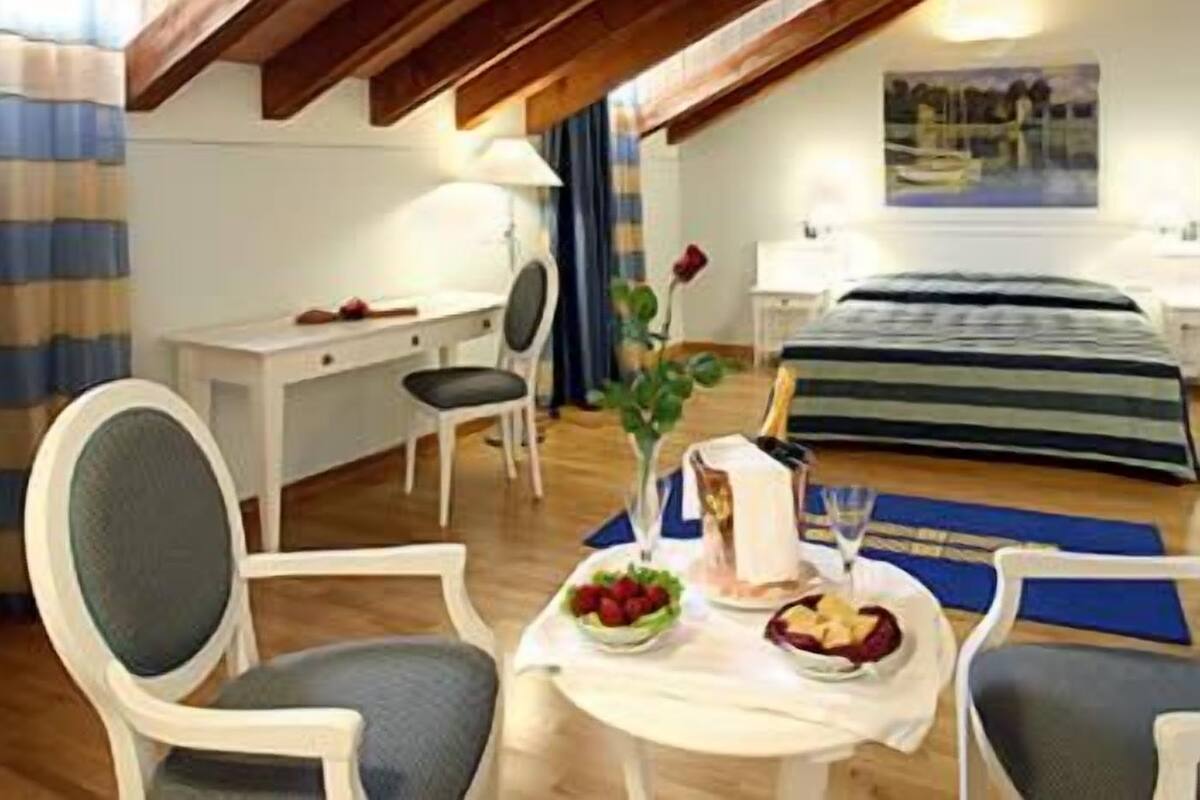 Une chambre confortable avec un lit, une table de chevet et un bureau avec une chaise. Il y a une fenêtre avec des rideaux, un tableau au mur et une table avec un vase et des fruits.