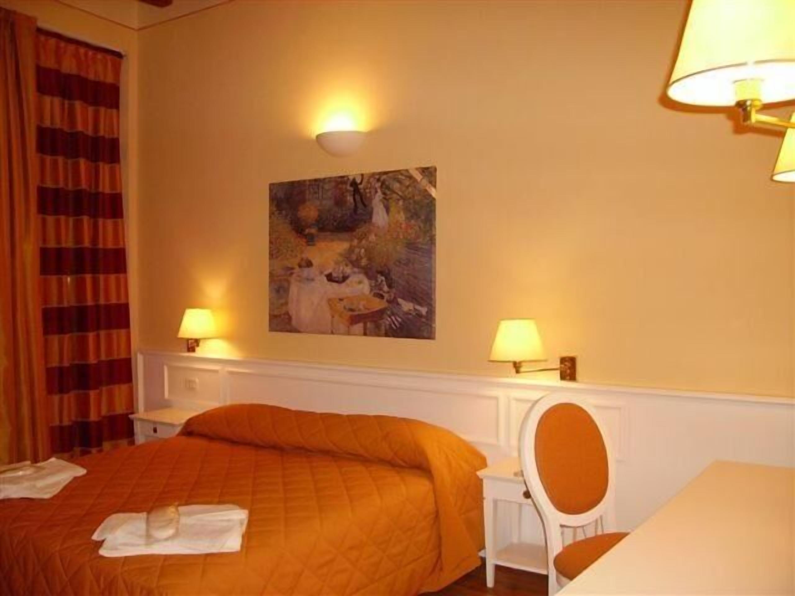 Habitación de hotel con cama, dos lámparas de noche, un cuadro en la pared, una silla y una mesa.