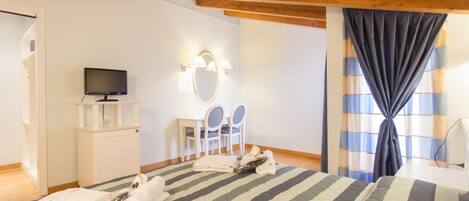 Un dormitorio con cama, televisor sobre un soporte, escritorio con sillas y ventana con cortinas.