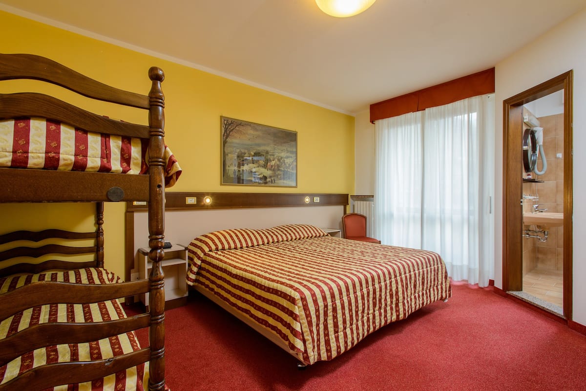 A hotel room with a double bed, a wooden headboard, a painting on the wall, a bathroom with a shower, and a red carpet.