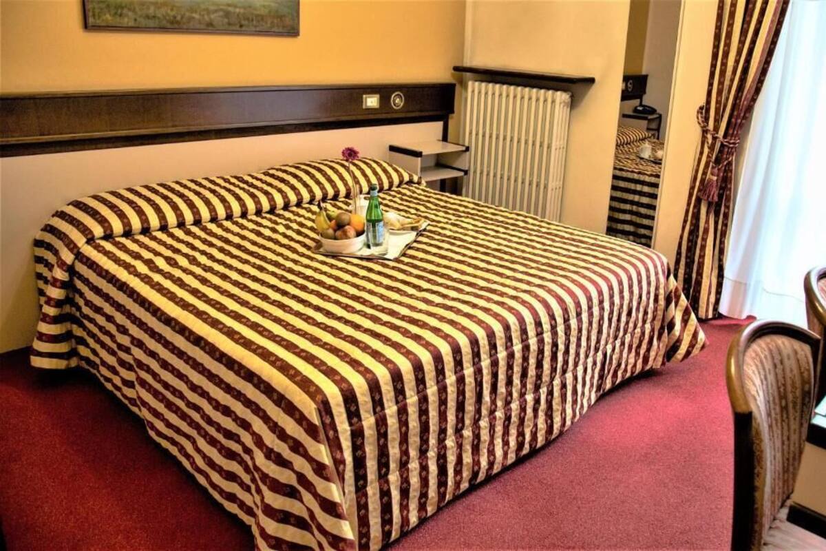 A hotel room with a striped bedspread, a bedside table with a lamp, and a small table with fruits and a bottle.