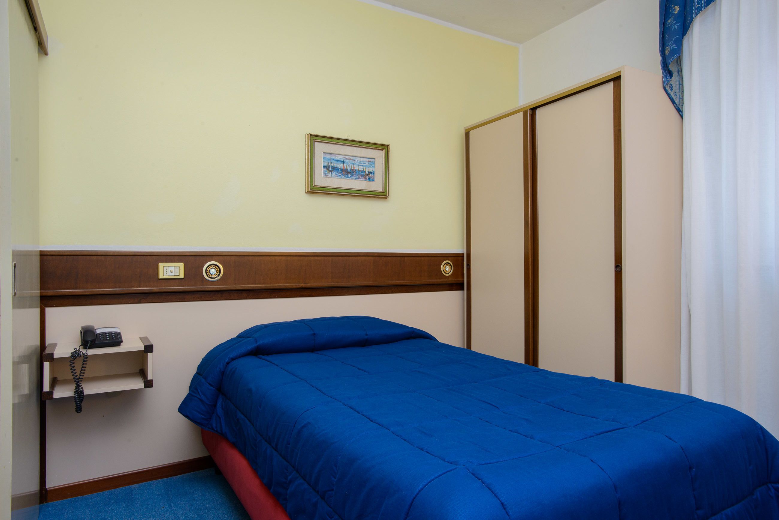 A hotel room with a single bed, a bedside telephone, a framed picture on the wall, and a wardrobe.