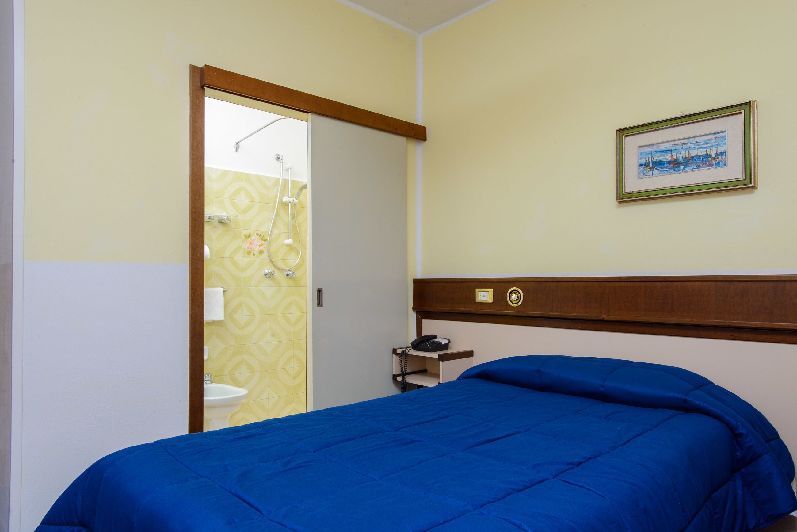 Quarto com cama, mesa de cabeceira, quadro na parede e banheiro visível através de uma porta aberta.
