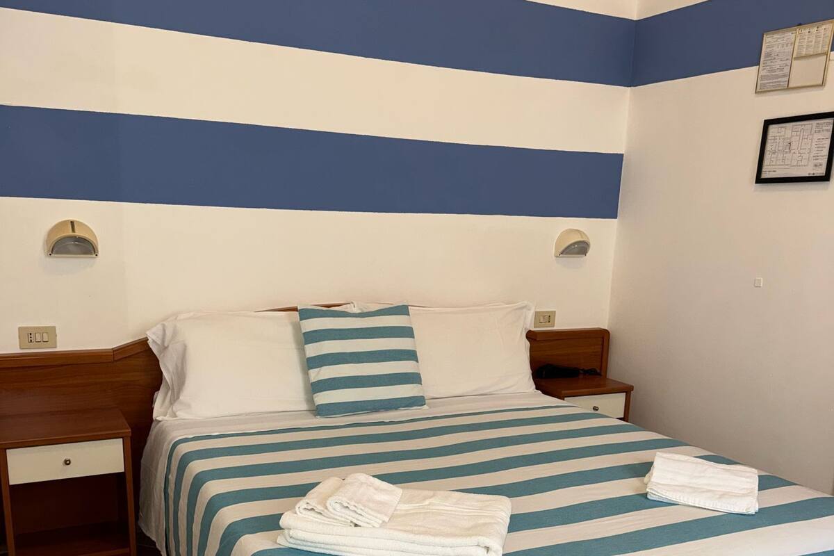 Hotellihuone, jossa on sängyssä sinivalkoraidallinen vuodevaatesetti, kaksi yöpöytää, puinen yöpöytä ja puinen tuoli.