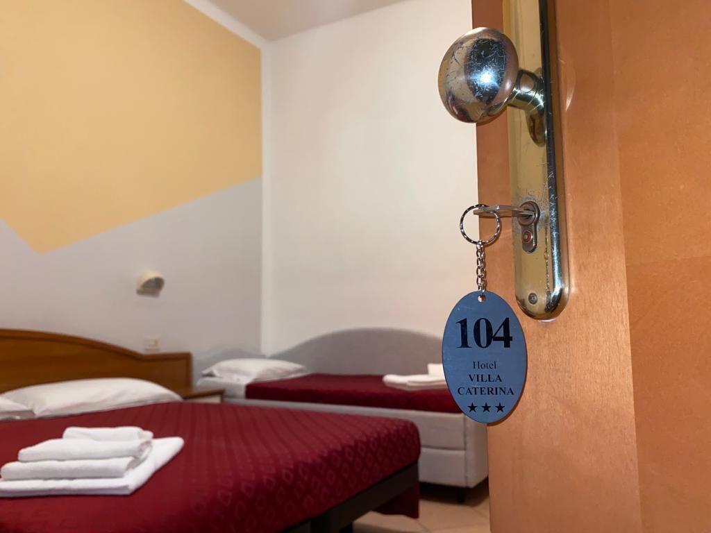 Habitación de hotel con dos camas, una con colcha roja y la otra con sábana blanca. Se ve un llavero con el número 104 y el nombre 'Hotel Villa Caterina' en el pomo de la puerta.