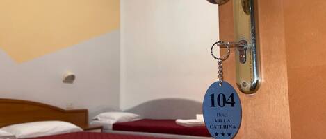 Habitación de hotel con dos camas, una con colcha roja y la otra con sábana blanca. Se ve un llavero con el número 104 y el nombre 'Hotel Villa Caterina' en el pomo de la puerta.