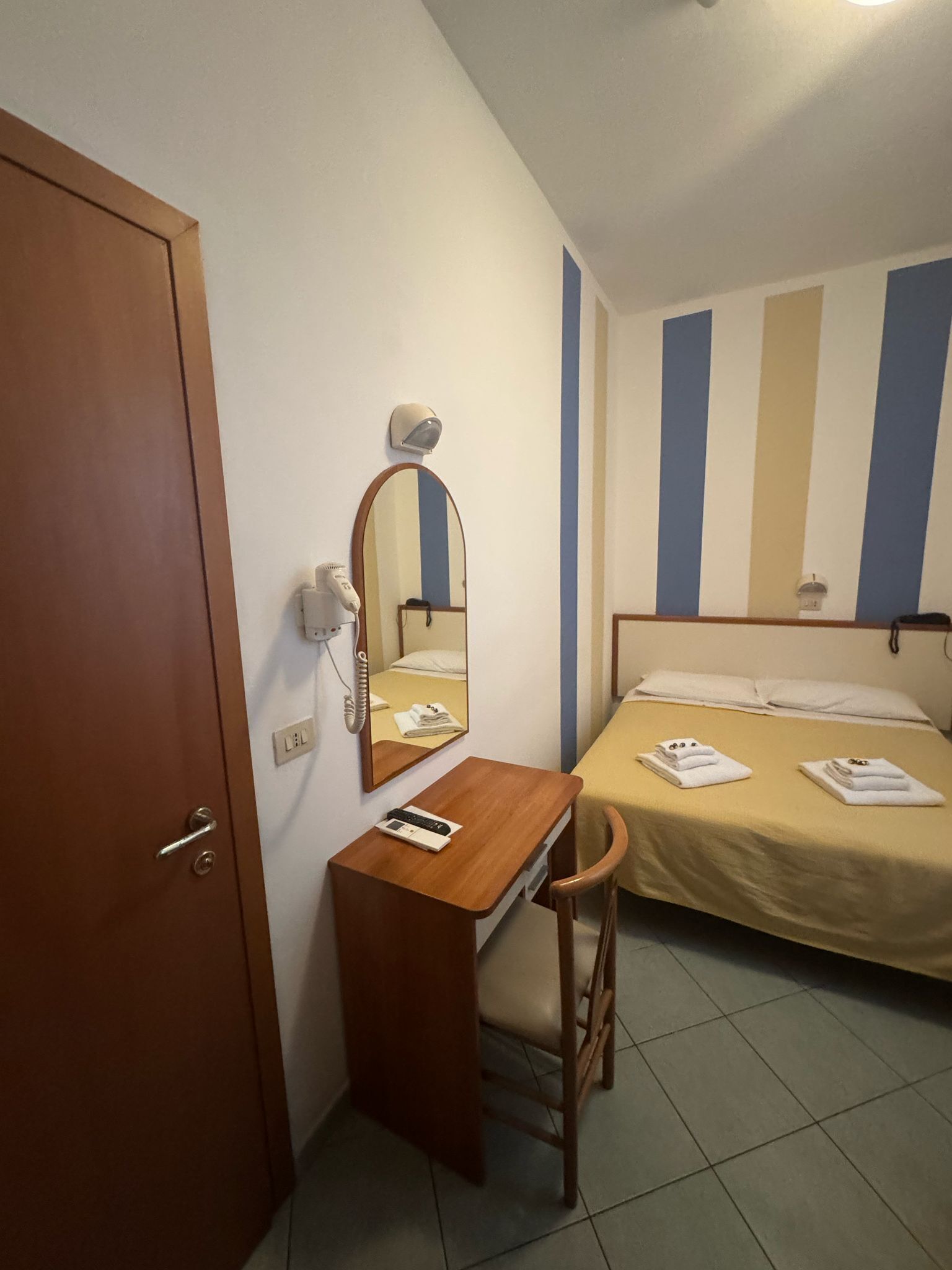 A small, single-room hotel room with a wooden desk, a chair, a bed with a striped wall, and a door.