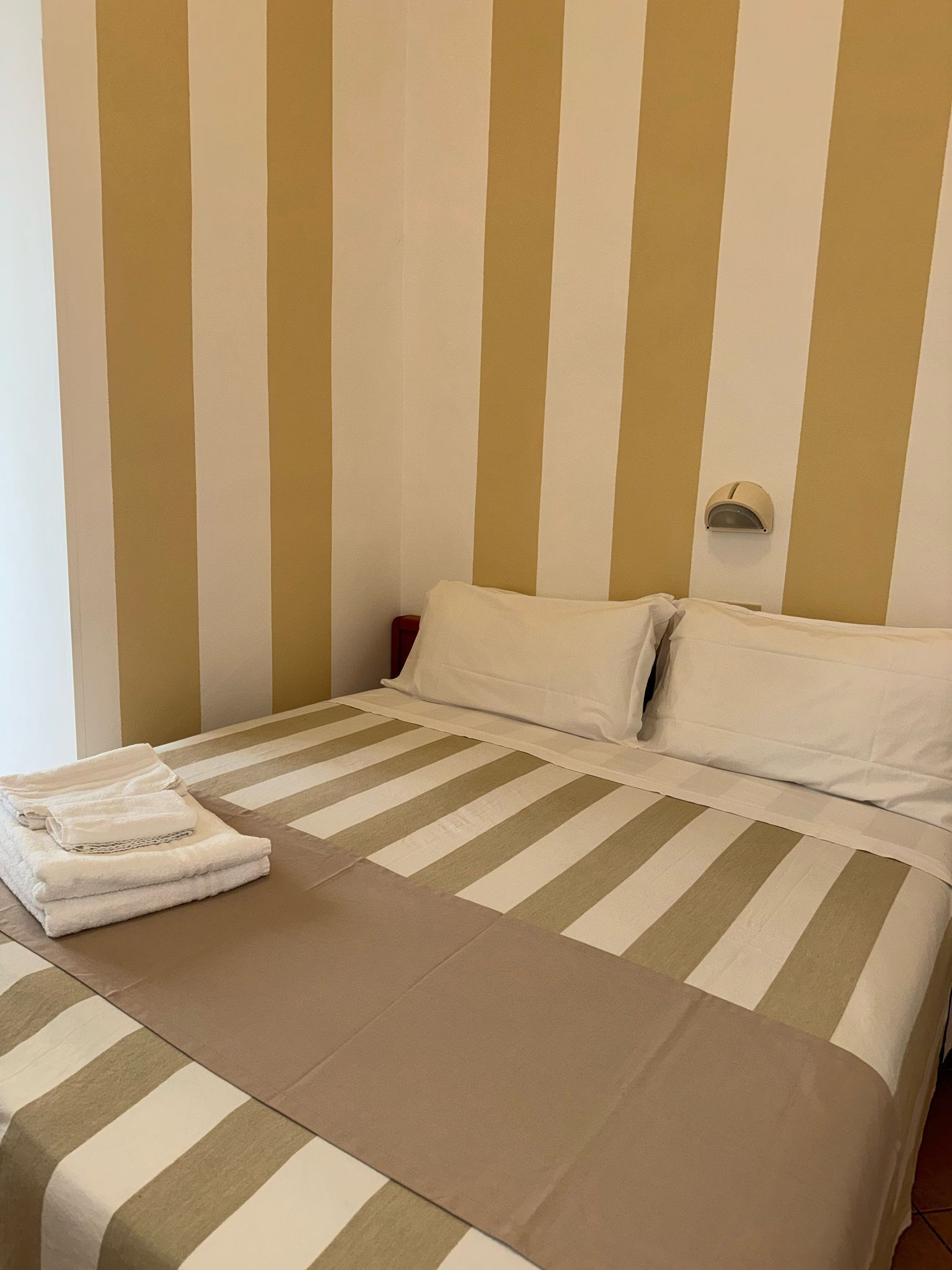 A hotel room with a bed featuring striped bedding in beige and white, a neatly folded white towel on the bed, and a wall-mounted night light.