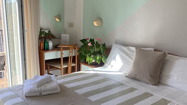 A neatly made bed with striped bedding, a desk with a chair, a potted plant, and a wall-mounted light fixture. - Hotel Villa Caterina (Rimini)