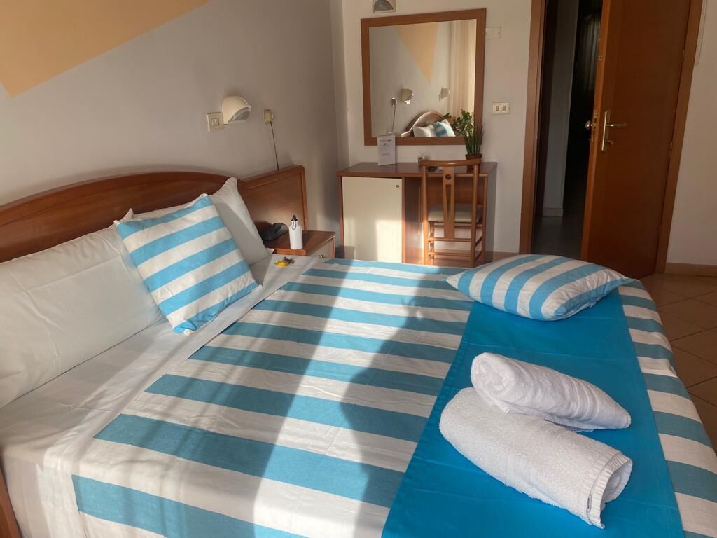 A hotel room with a bed featuring blue and white striped bedding, a wooden headboard, a bedside table with a lamp, a small desk with a mirror, and a door leading to another room.