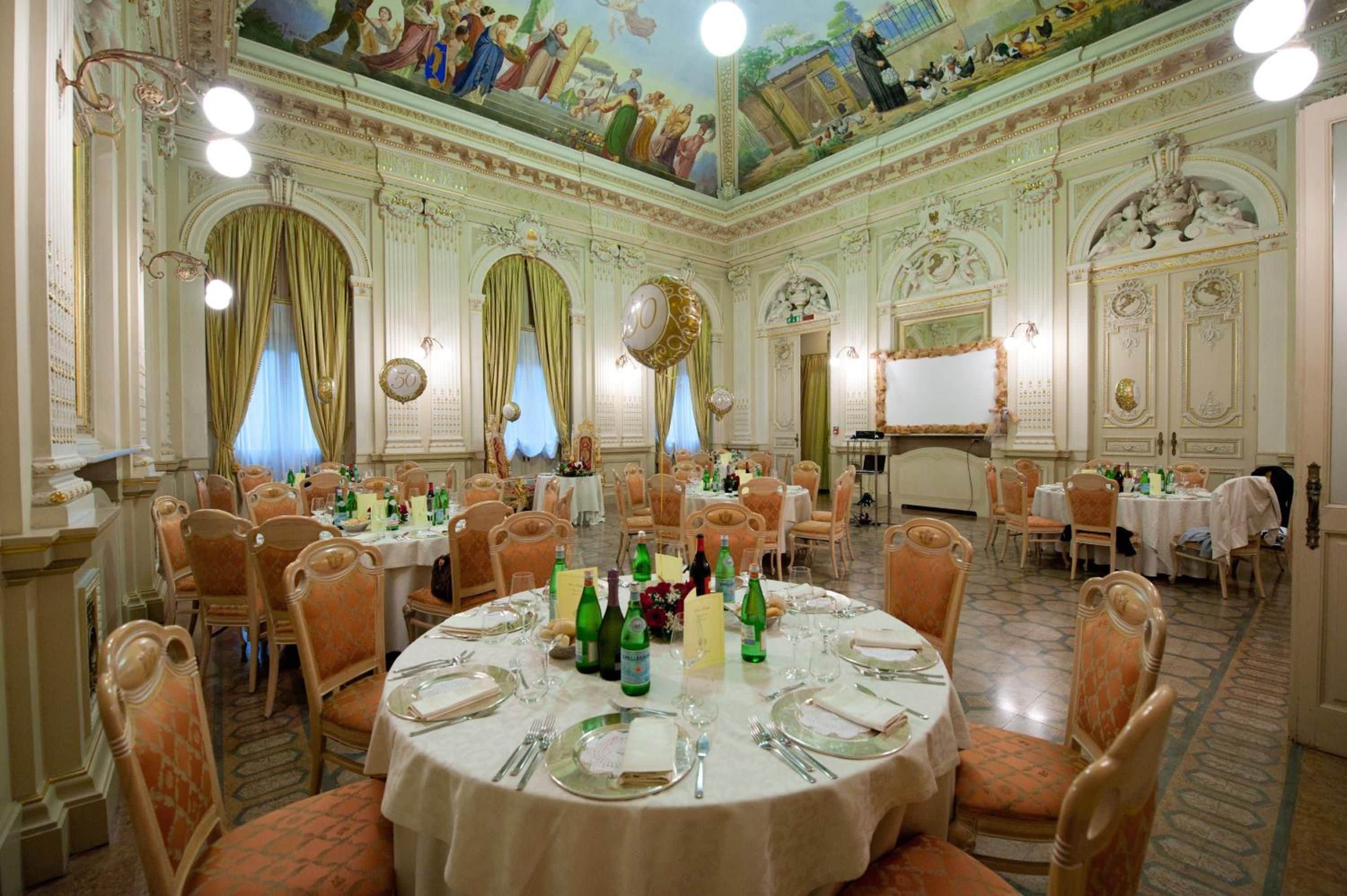 Un salón de banquetes con mesas redondas dispuestas para cenar, frescos ornamentales en el techo y detalles arquitectónicos clásicos.