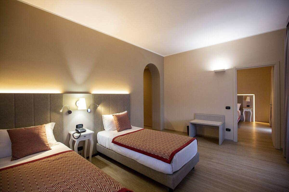 A hotel room with two beds, a bedside table with a phone, a lamp, and a bench.