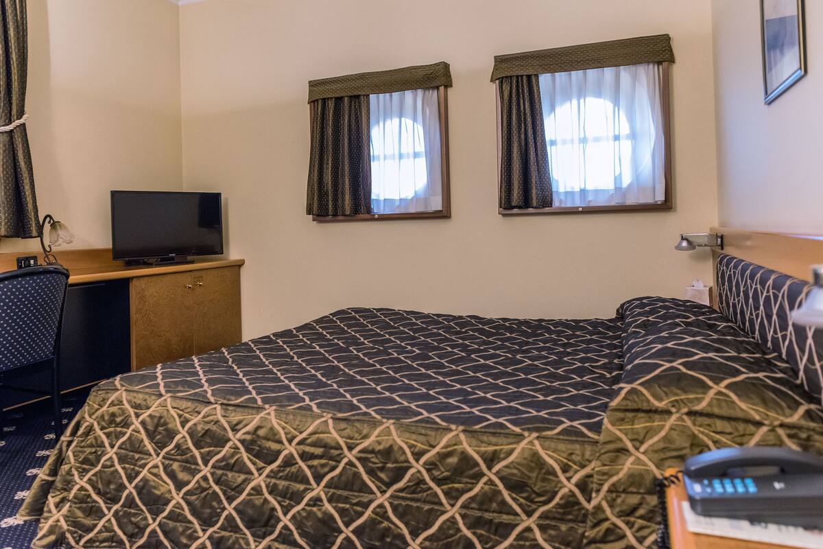A hotel room with a bed, a desk with a TV, two windows with curtains, and a phone on the bedside table.