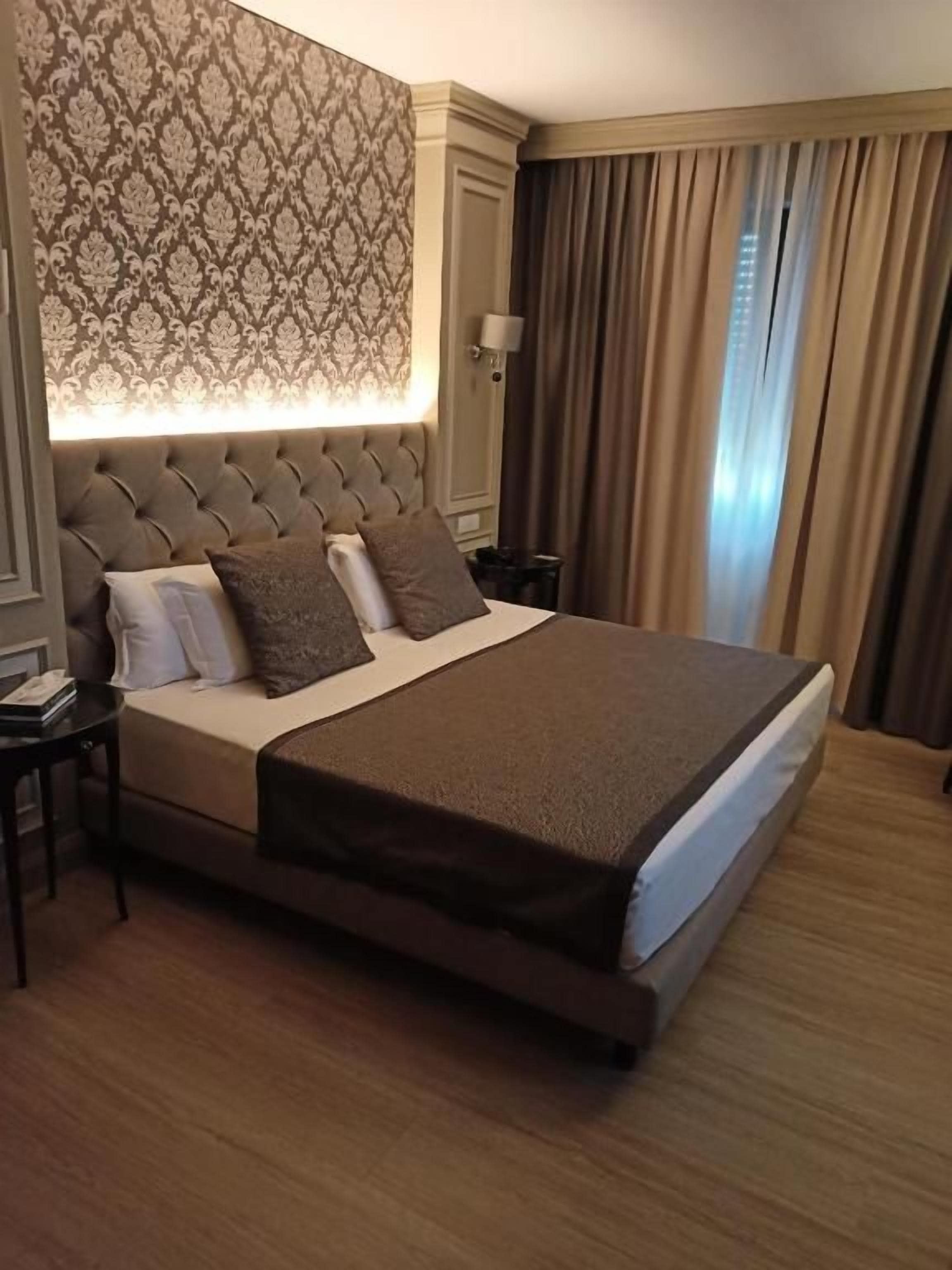 Habitación de hotel con una cama grande, cabecera con panel iluminado, mesita de noche y ventana con cortinas.
