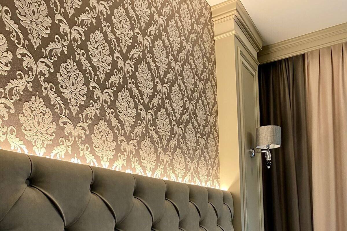 A hotel room with a tufted headboard, a patterned wallpaper, and a bed with a quilted mattress and two pillows.