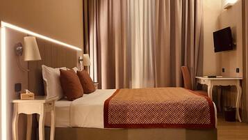 Habitación de hotel con cama, mesitas de noche, escritorio, televisor y un gran ventanal con cortinas.