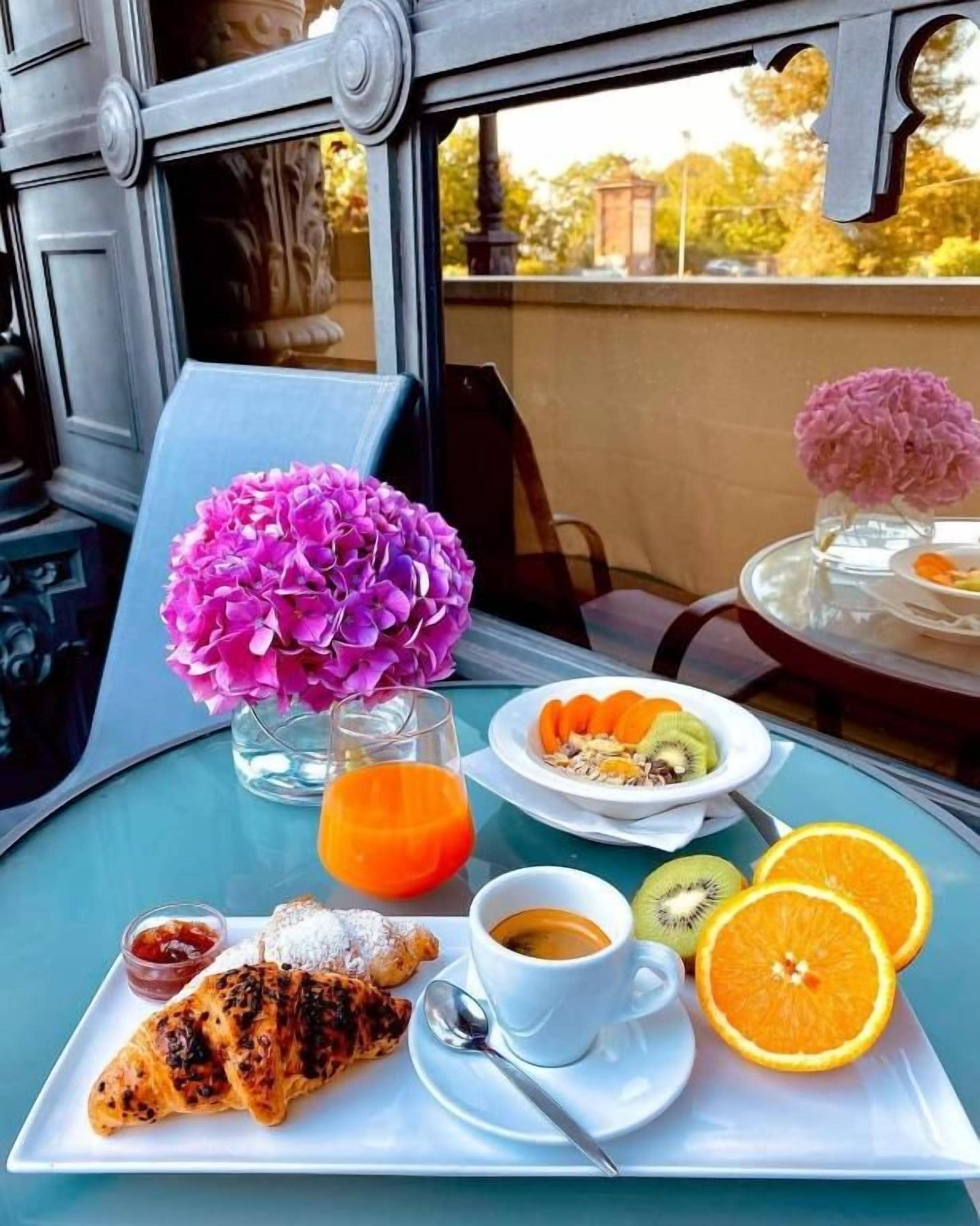 A breakfast setting with a croissant, orange slices, kiwi, coffee, and a vase of flowers on a table.