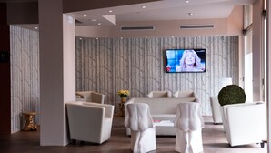 A modern living room with a large flat-screen TV mounted on the wall, a sofa, and two unique white chairs. - Hotel Oro Blu (Milan)