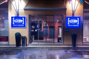 A hotel entrance with glass doors, a reception desk, and two large blue signs indicating the hotel name and its proximity to local attractions.