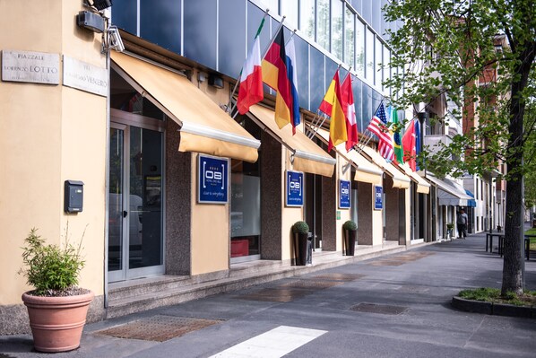A hotel facade with multiple flags, a clear entrance, and a sign indicating amenities. - Hotel Oro Blu (Milan)