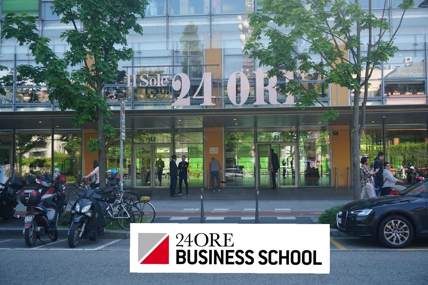 Exterior de una escuela de negocios abierta las 24 horas, con fachada de vidrio, motocicletas estacionadas y personas entrando y saliendo.