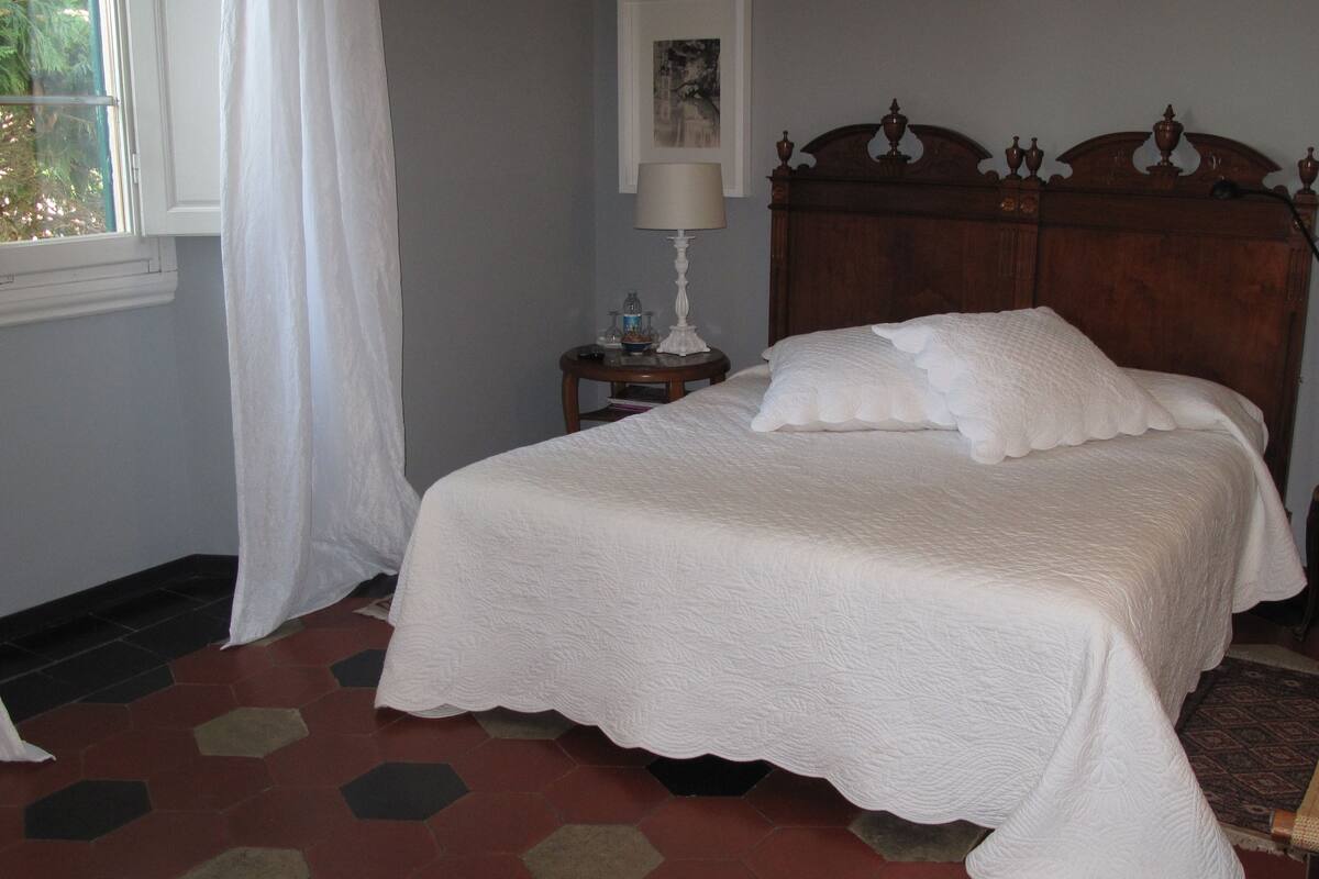 Un lit bien fait, avec du linge de lit blanc, une tête de lit en bois et une petite table de chevet sur laquelle se trouve une lampe.