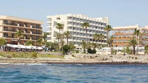 Un hotel costero con varios pisos, palmeras y servicios junto a la playa.