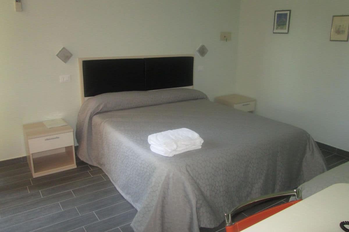 Un lit bien fait, recouvert d’une couverture grise et agrémenté d’une serviette blanche pliée, une table de chevet avec une lampe et un interrupteur fixé au mur.