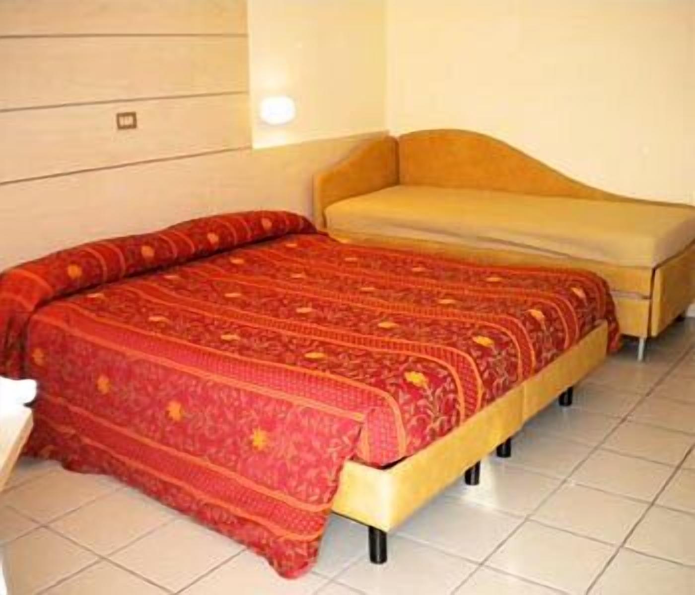 A bed with a red patterned bedspread and a yellow headboard in a room with white tiled flooring.