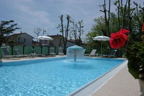 A swimming pool with a water fountain, lounge chairs, and umbrellas. - Hotel Prater (Cesenatico)