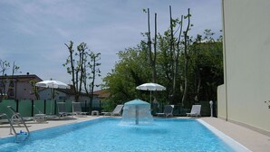 A swimming pool with a water fountain, surrounded by lounge chairs and umbrellas. - Hotel Prater (Cesenatico)
