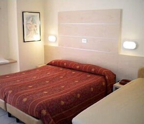 A single bed with a patterned bedspread, a nightstand with a lamp, and a framed picture on the wall. - Hotel Prater (Cesenatico)