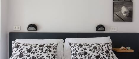 Una cama bien tendida con almohadillas de estampado floral, un cabecero oscuro y un pequeño estante de madera con adornos.
