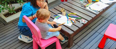 A child drawing at an outdoor wooden table with a pink chair, surrounded by art supplies.
