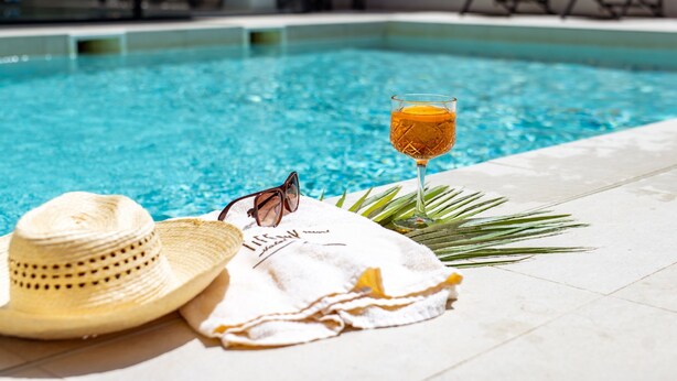 Escena junto a la piscina con un vaso de jugo de naranja, un sombrero de paja, gafas de sol y una toalla.