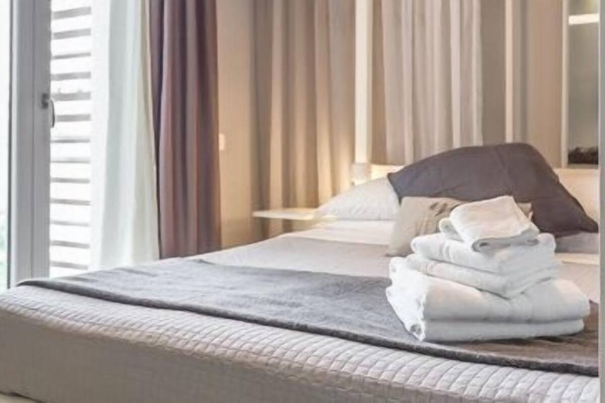 A neatly made bed with a grey blanket and white towels, a bedside lamp, and a window with curtains.