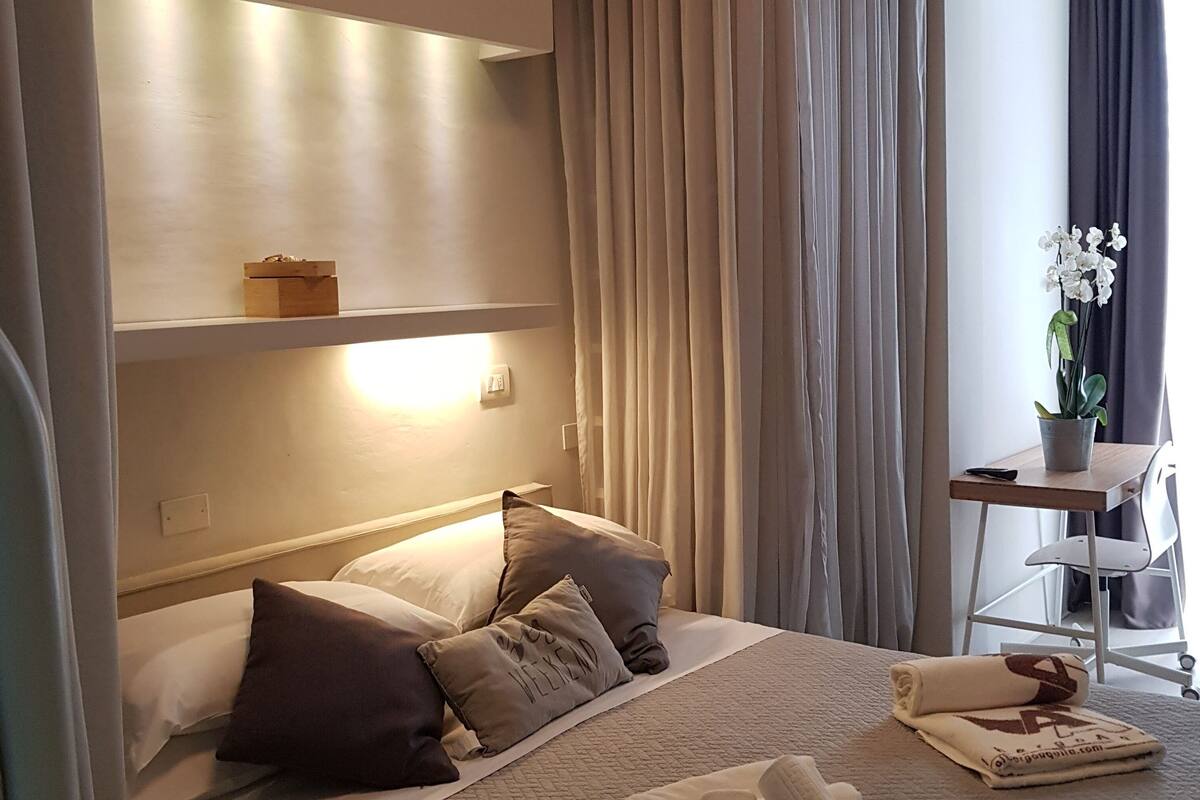 A hotel room with a bed, pillows, a bedside table with a lamp, a chair, and a window with curtains.