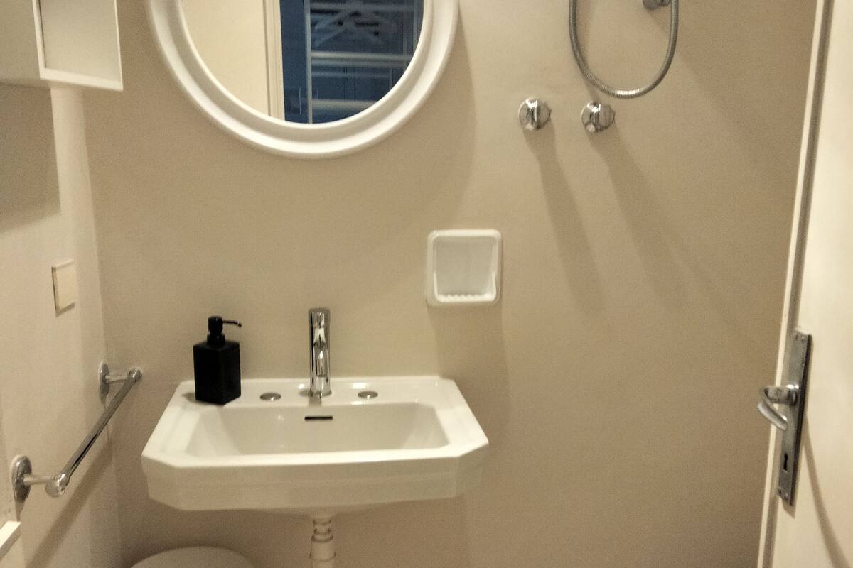 A small bathroom with a white sink, a mirror, a shower, and a wooden bench.