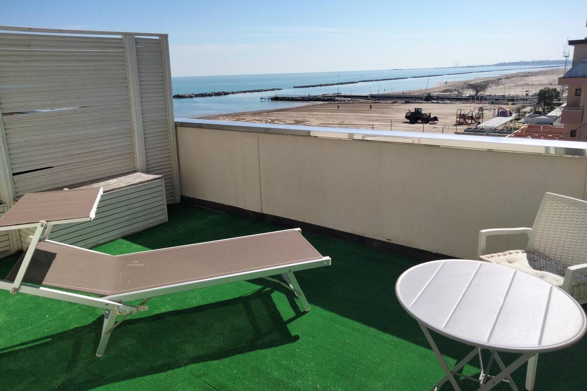 A balcony with green artificial turf, two lounge chairs, and a small table. A view of the beach and sea is visible through the railing.