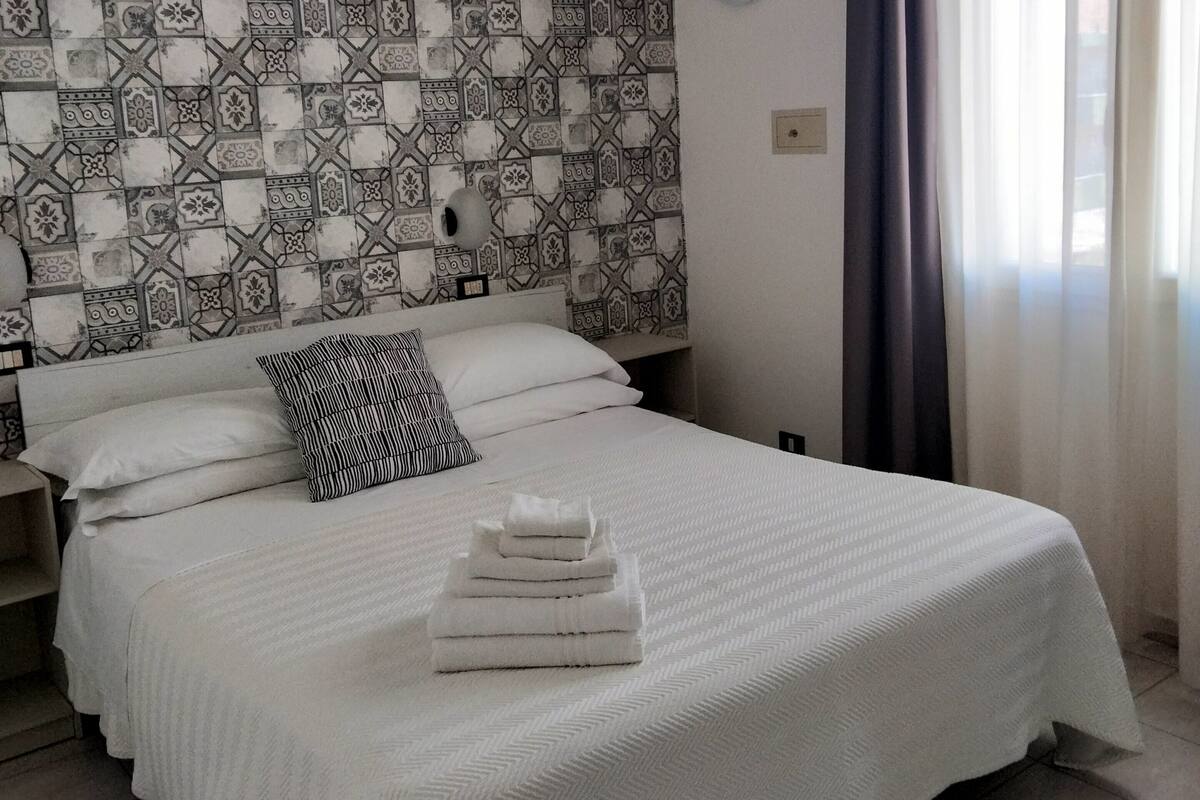 A neatly made bed with white linens and a stack of folded towels, a patterned tile wall, a round wall clock, and a window with sheer curtains.