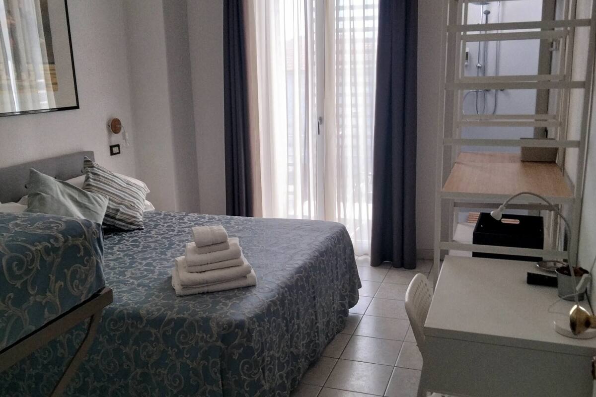A hotel room with a bed, a desk, a chair, a hanging rack, and a window with curtains.