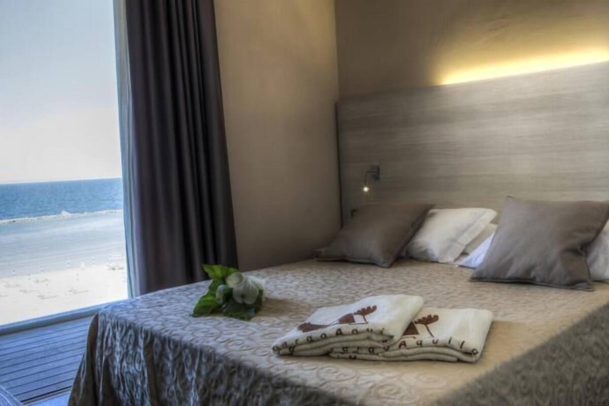 A hotel room with a bed, pillows, a bedside table, and a view of the beach.