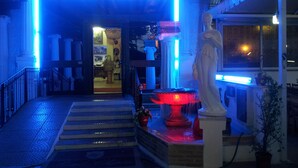 A hotel entrance with a neon sign, a statue, a fountain, and a staircase leading inside.