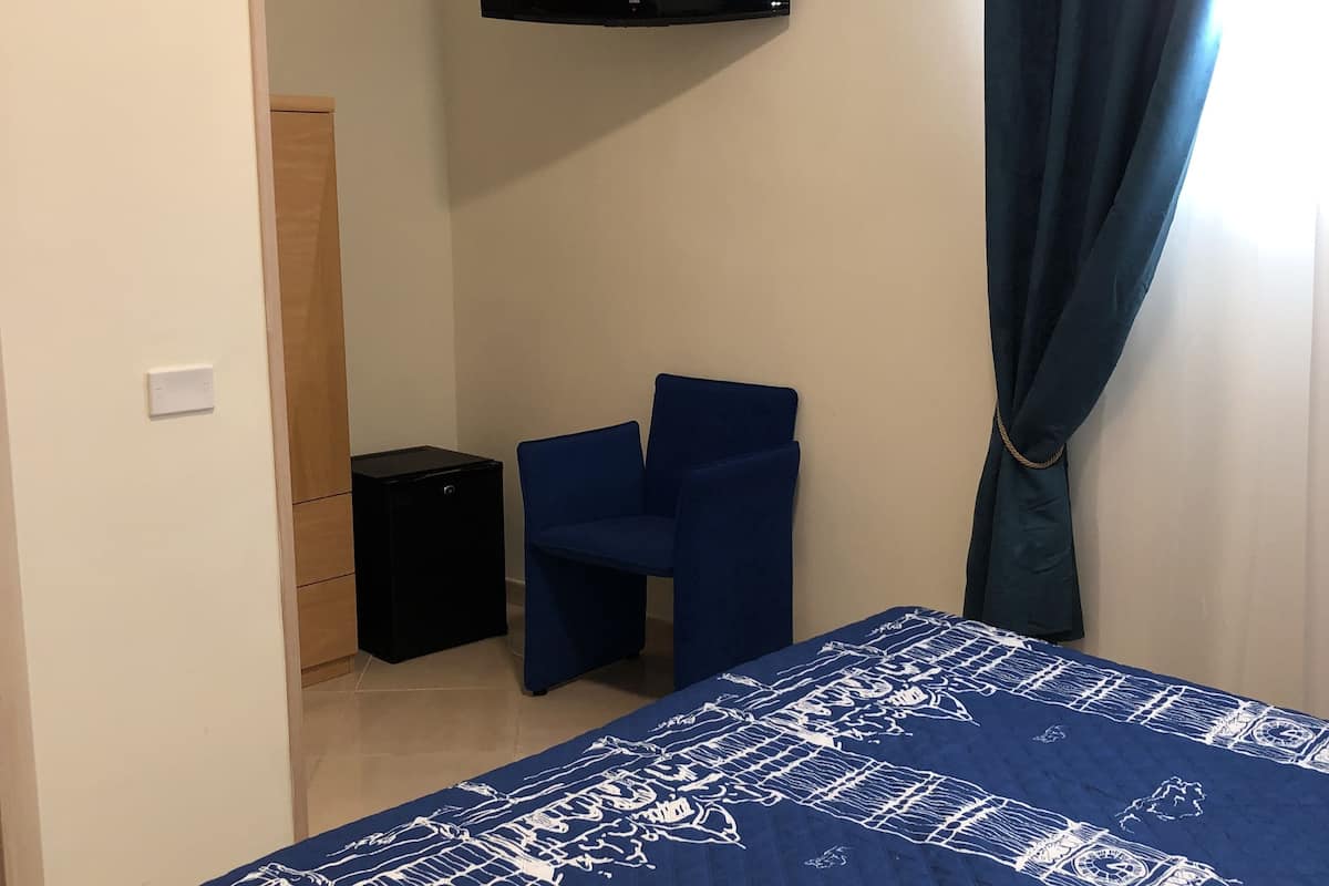 Ein Schlafzimmer mit einem Bett, einem flachbildfernseher an der Wand, einem kleinen schwarzen Lautsprecher und einem blauen Stuhl.
