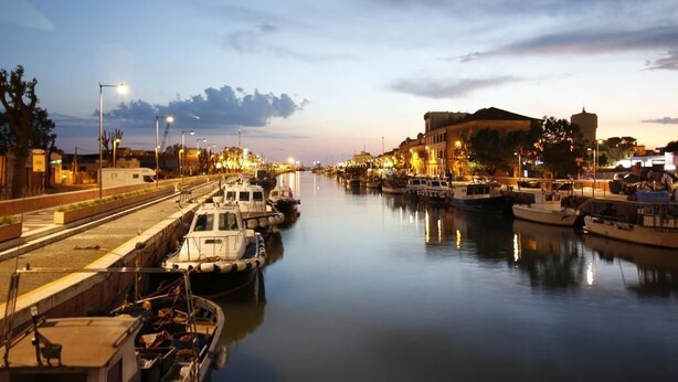A marina with boats docked, a calm river, and buildings along the riverbank.