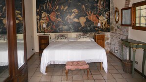 A bedroom with a large tapestry, a bed with a white bedspread, a wooden bench, and a stone wall.