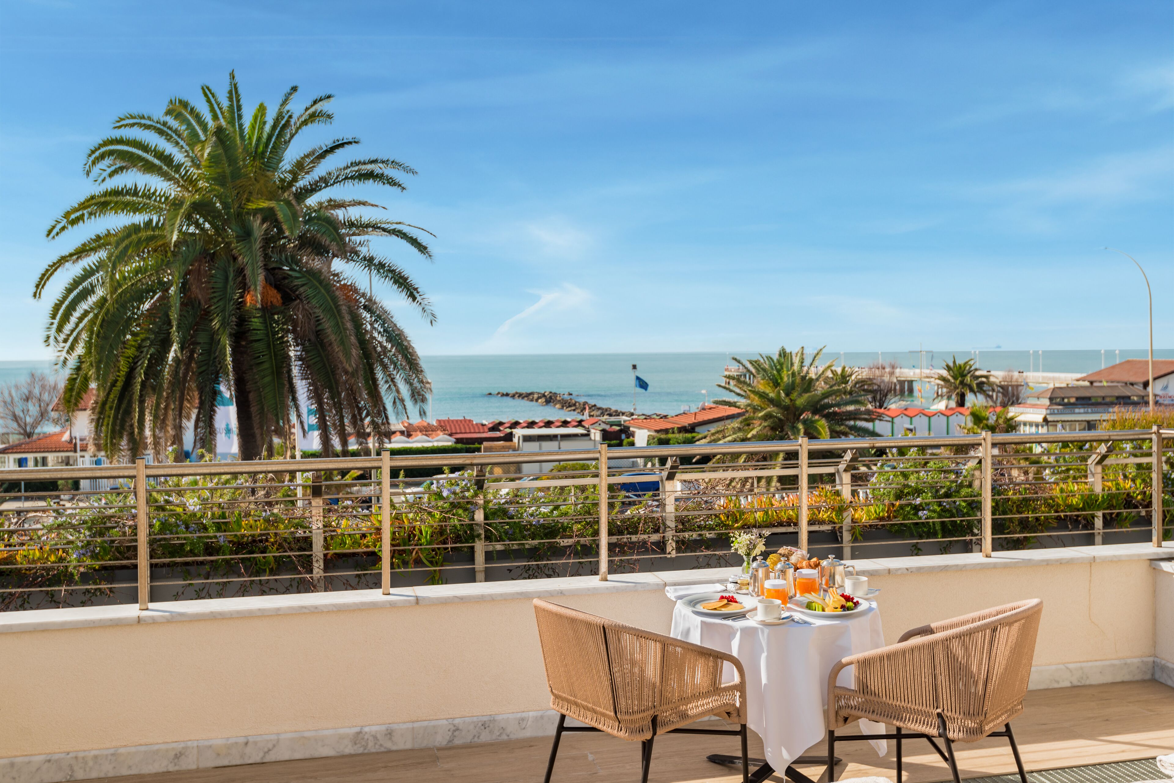 Ein Balkon mit Frühstückstisch, mit Blick auf einen Strand mit Palmen und wolkenlosen Himmel.