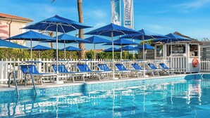 Una piscina con sombrillas azules y sillas de descanso, palmeras y una bandera con texto.