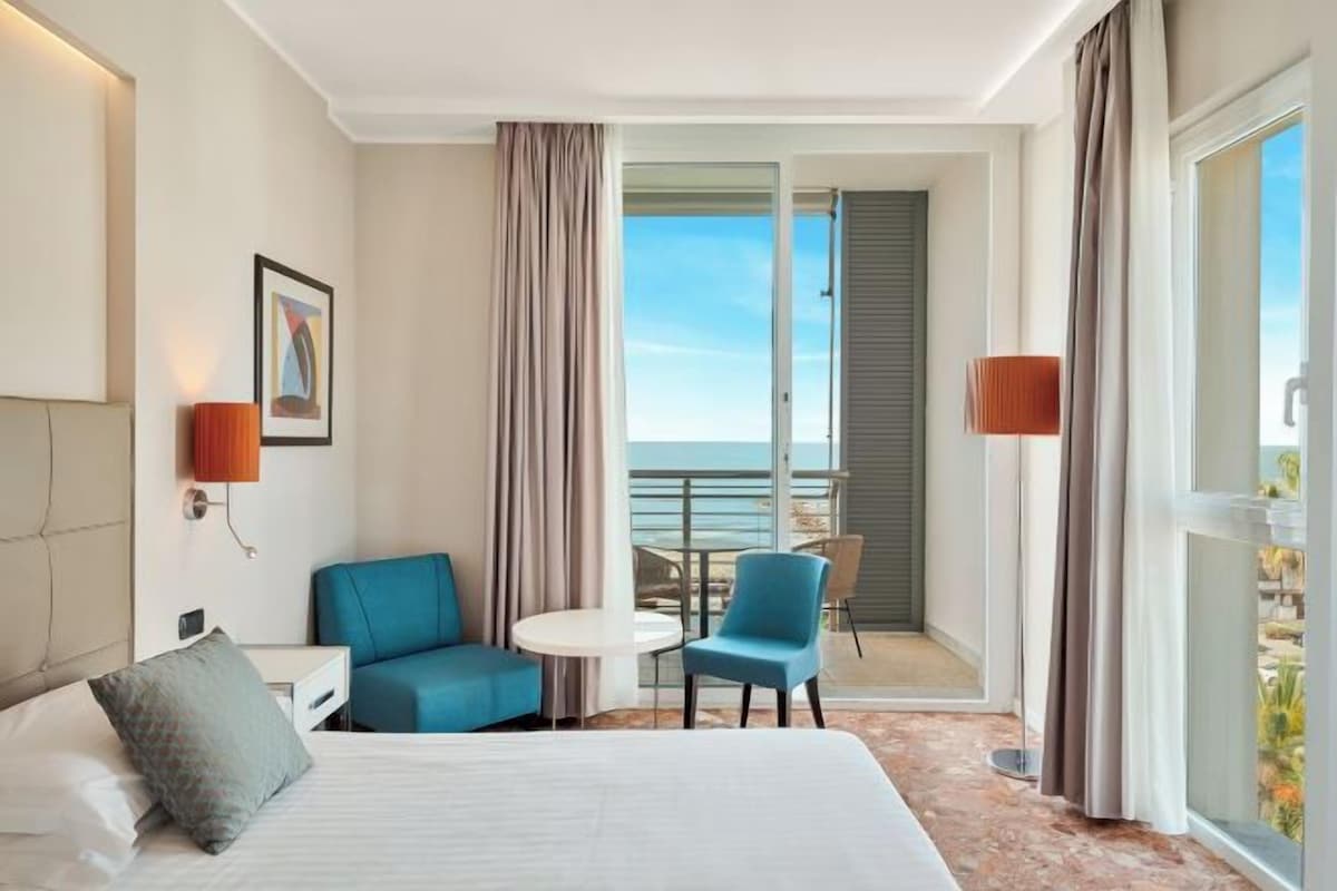 A hotel room with a bed, a small table, two chairs, a balcony with a view of the sea, and a painting on the wall.